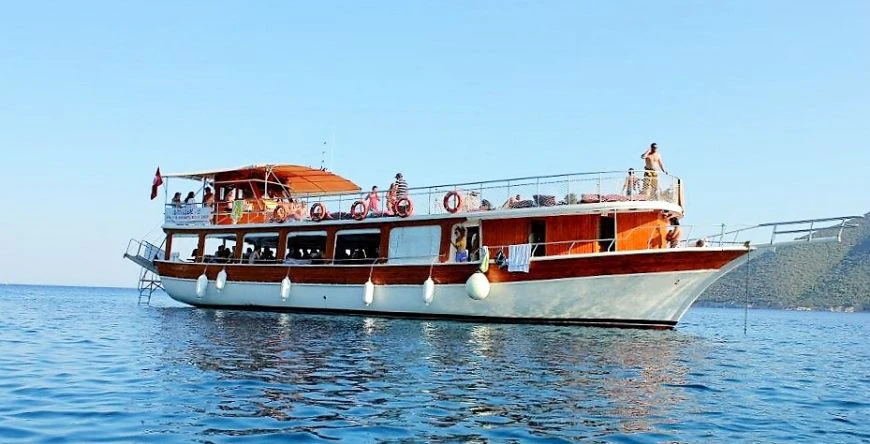 A wooden boat with people on the deck sailing on calm water under a clear blue sky.