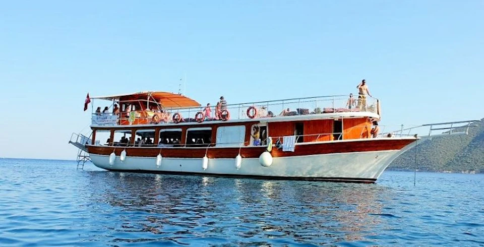 A wooden boat with people on the deck sailing on calm water under a clear blue sky.
