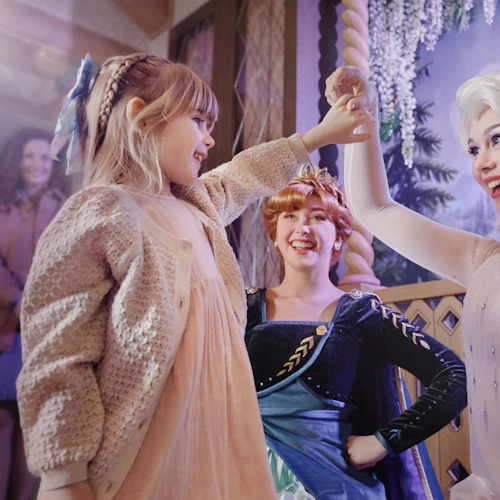 A child holding hands with performers dressed as fairytale characters in a themed setting with a smiling onlooker.