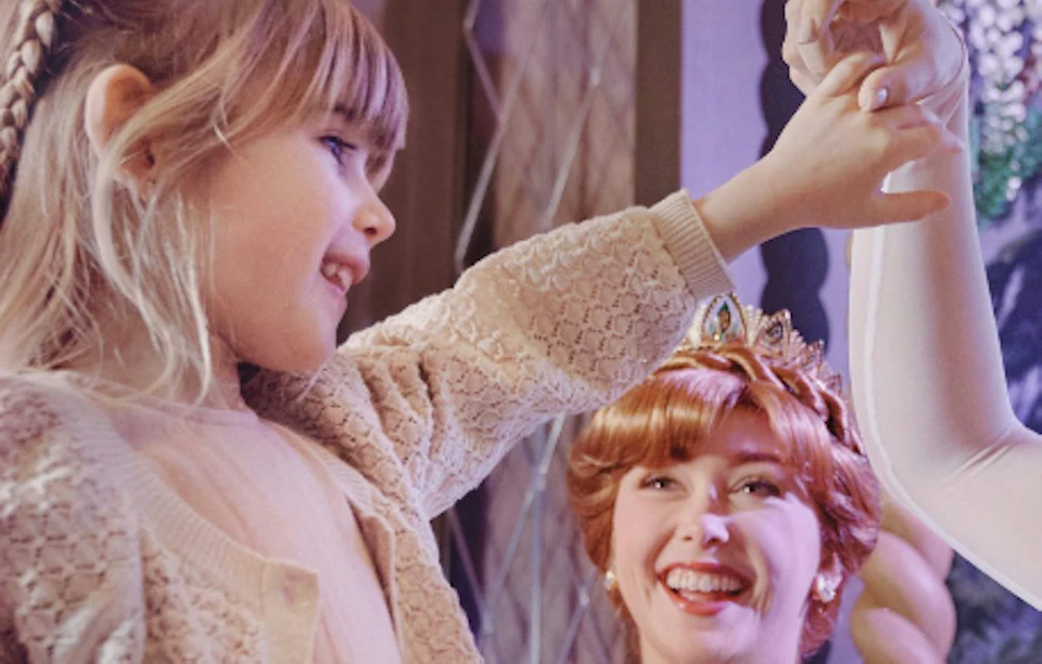 A child holding hands with performers dressed as fairytale characters in a themed setting with a smiling onlooker.