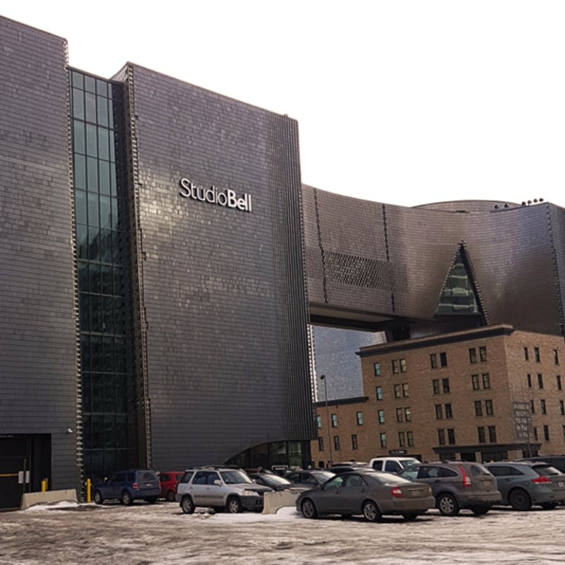 Moderne bygning mærket "Studio Bell" med et unikt design, tilstødende ældre struktur og snedækkede parkerede biler foran.