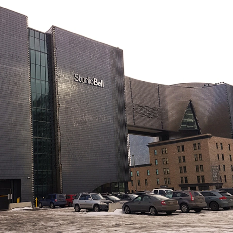 Modern building marked "Studio Bell" with a unique design, adjacent older structure, and snow-dusted parked cars in front.