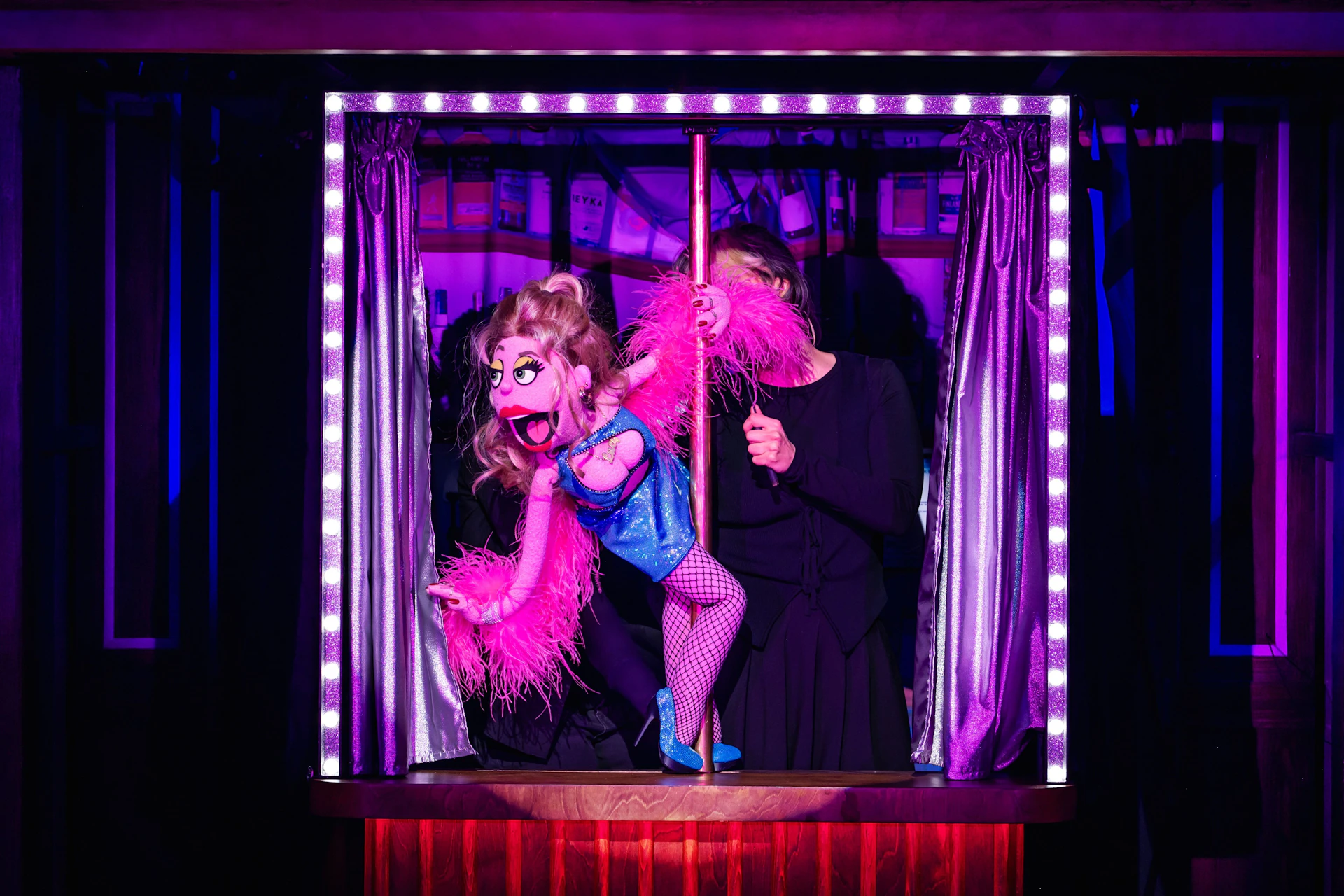 A colorful puppet in a blue outfit and pink feather boa is operated by a person dressed in black on a small stage with curtains.
