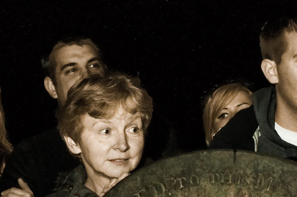 A group of people stand around old tombstones in a dimly lit graveyard, one person with face paint resembling a wound.