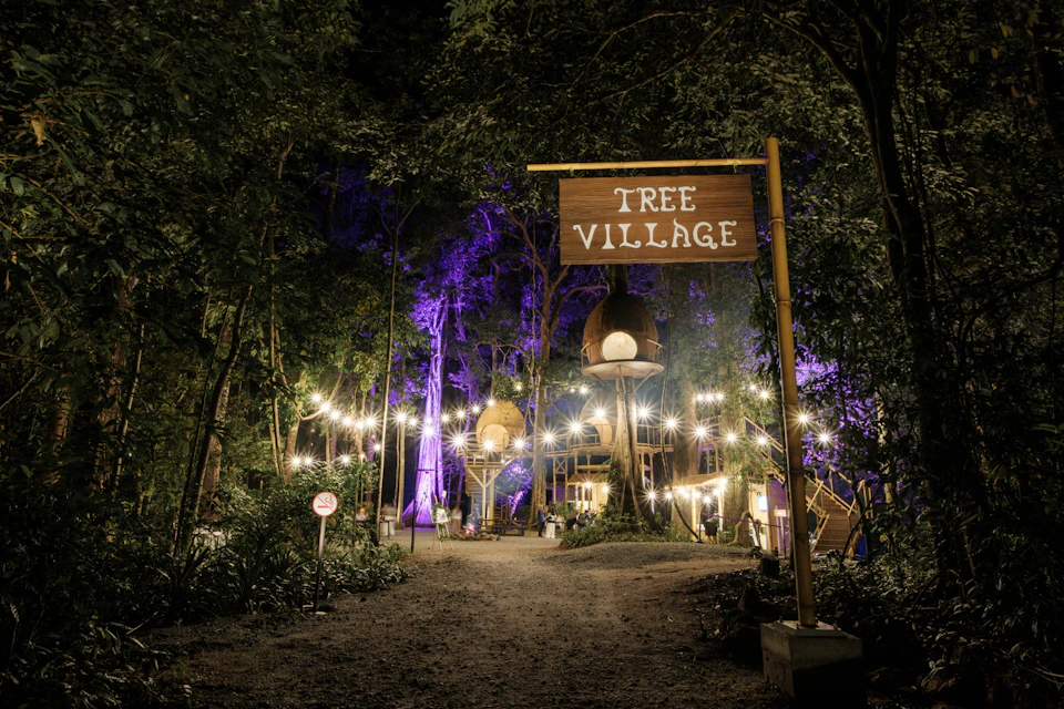 A nighttime scene of a forest setting with treehouses lit by string lights and a wooden sign reading 'Tree Village.'