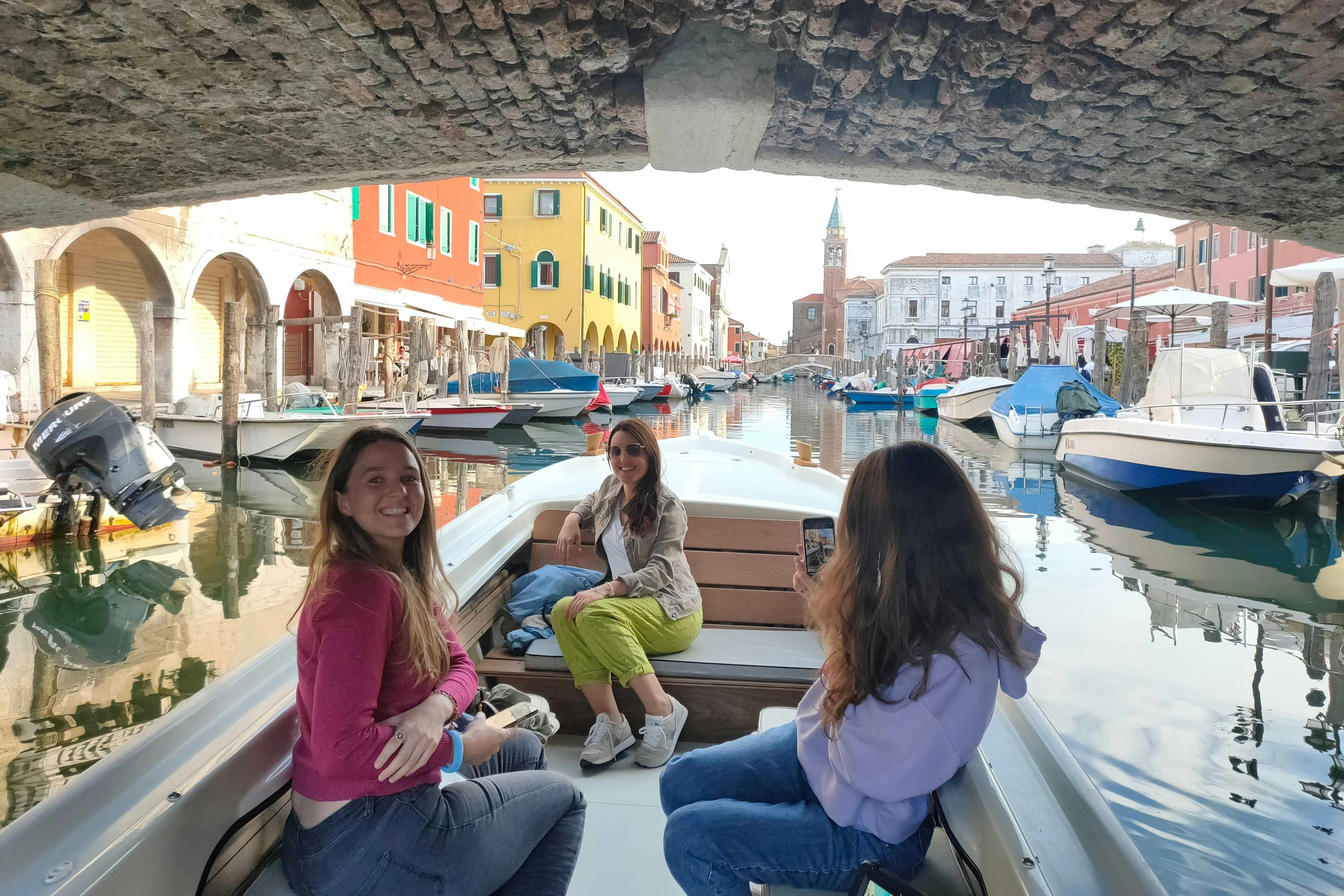 Three people on a boat under a brick bridge in a colorful canal with buildings and boats lining the waterway.