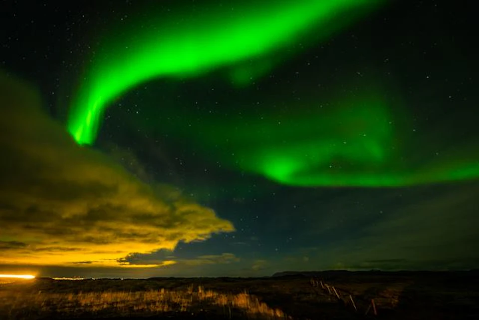 Green aurora borealis lights up the night sky over a dark landscape with clouds and stars visible.