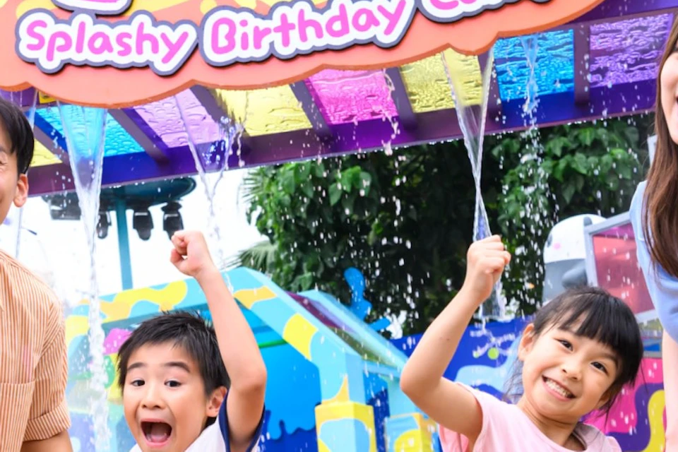 A family of four joyfully posing in front of a colorful amusement park entrance with a sign reading "Panda Friends Splashy Birthday Camp."