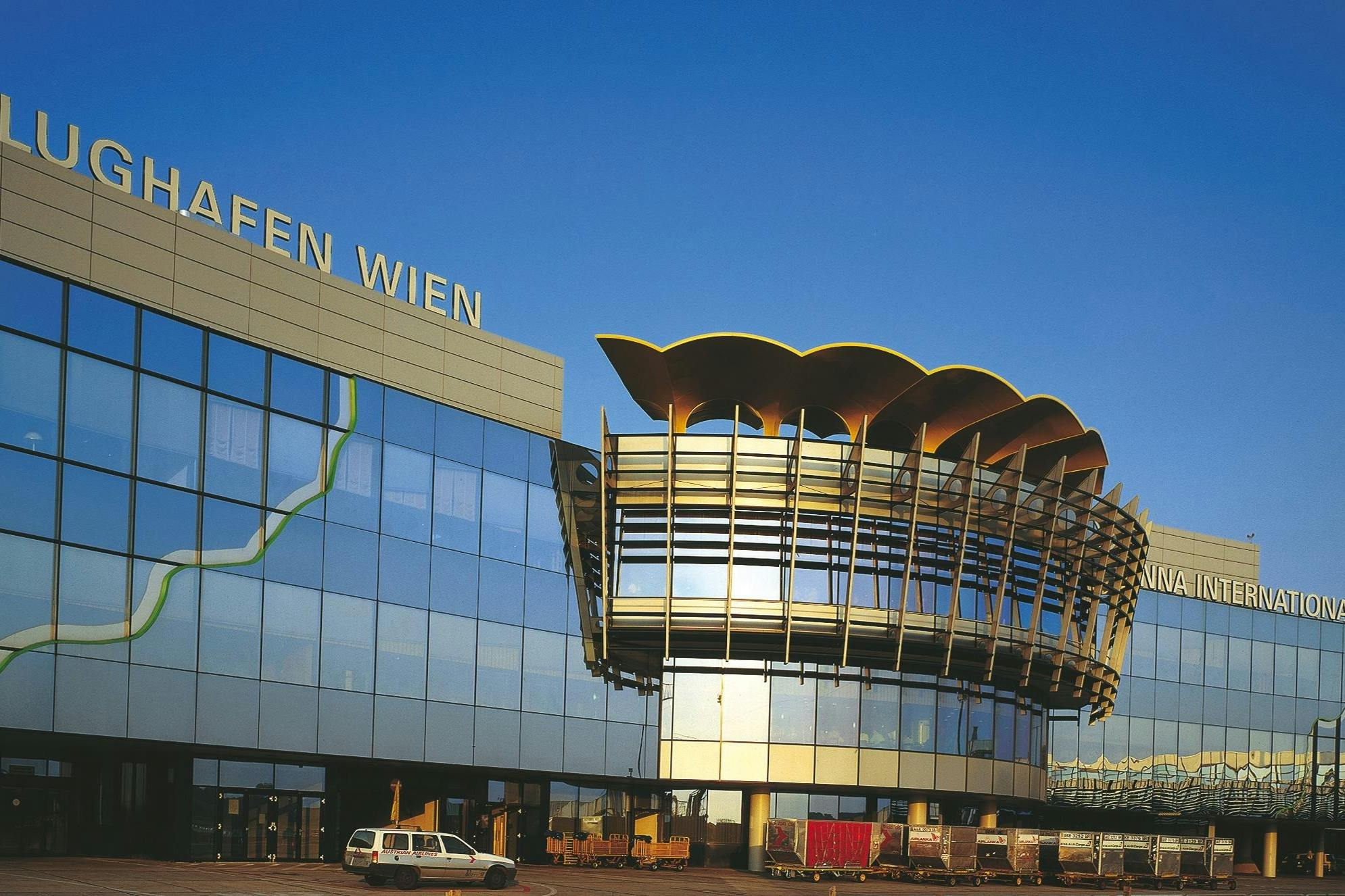 一栋现代建筑的玻璃立面，文字为“FLUGHAFEN WIEN” ，建筑结构独特；停放着车辆和行李车。