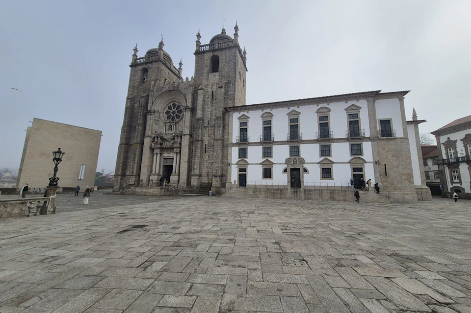 A large historic stone cathedral with two towers and a rose window adjacent to a white building in a cobblestone square.