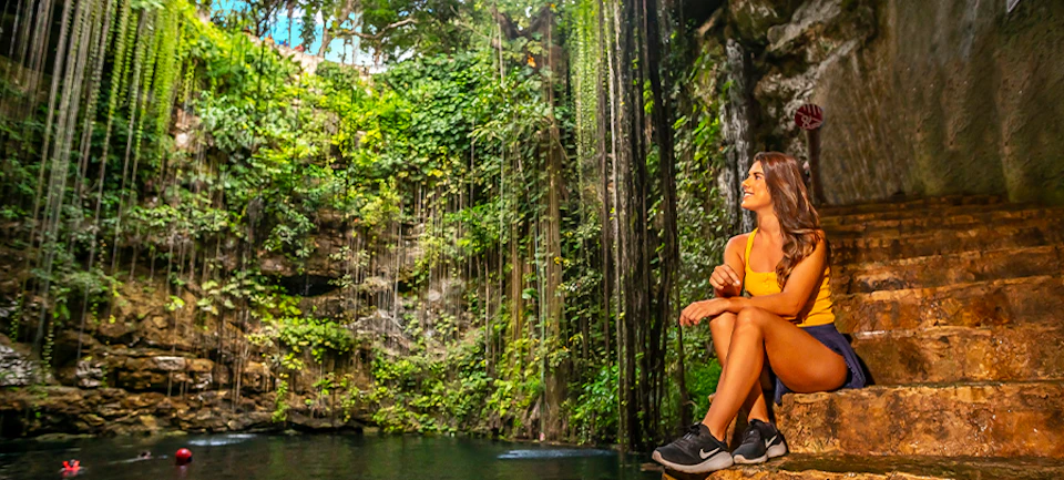 Woman in athletic wear sits on ledge near a lush, green cenote with hanging vines and rock formations.