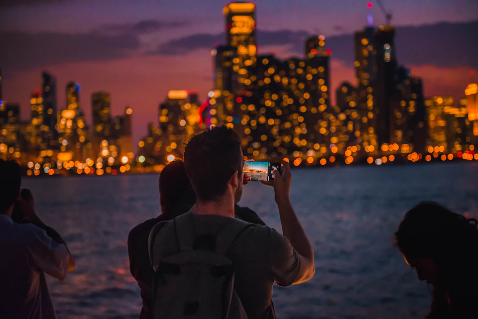 A person with a backpack photographs a city skyline illuminated with lights at dusk, viewed from across a body of water.