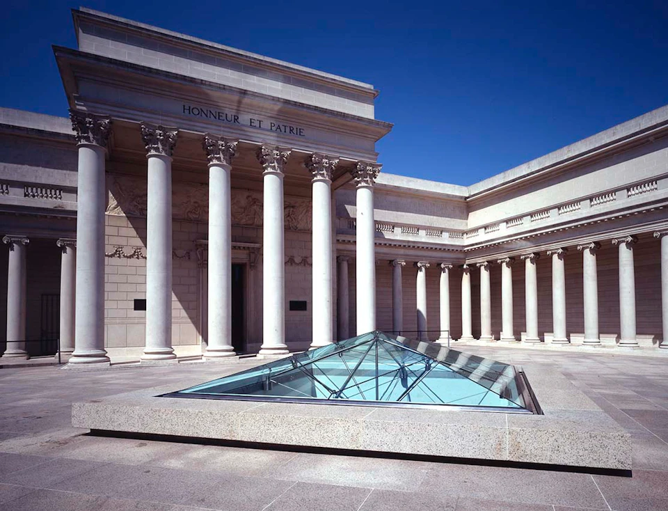 Neoclassical building with tall columns and a courtyard featuring a glass pyramid skylight under a clear blue sky.
