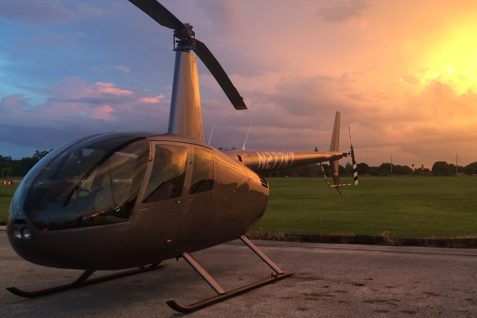 A parked helicopter on a concrete surface with a grassy field and a vibrant sunset sky in the background.