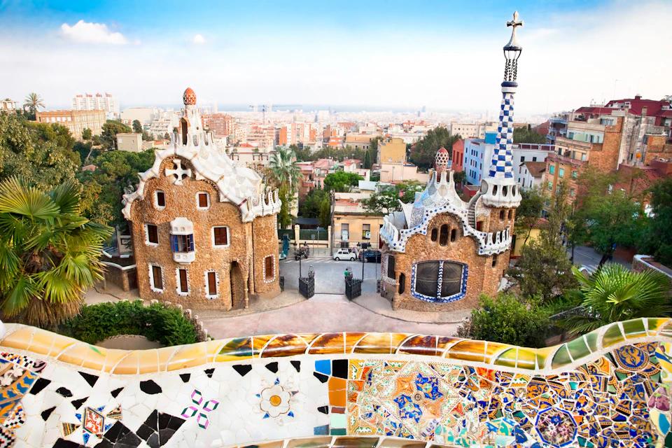 Colorful, mosaic-tiled wall with whimsical, Gaudí-designed buildings and cityscape in the background at Park Güell, Barcelona.
