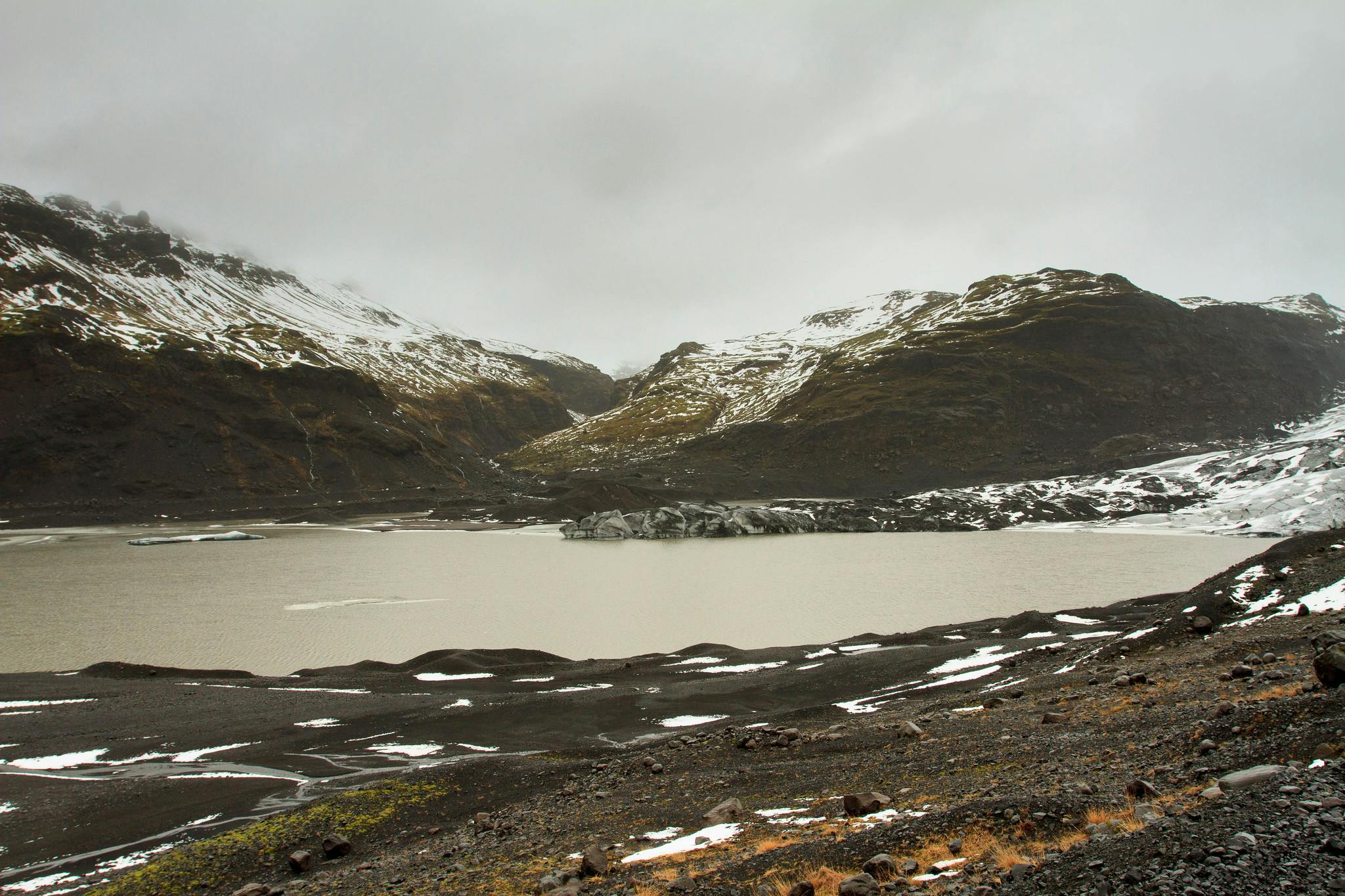 白雪皑皑的山脉环绕着泥泞的湖泊，在多云的天空下，前景中有一片雪地和岩石地形。