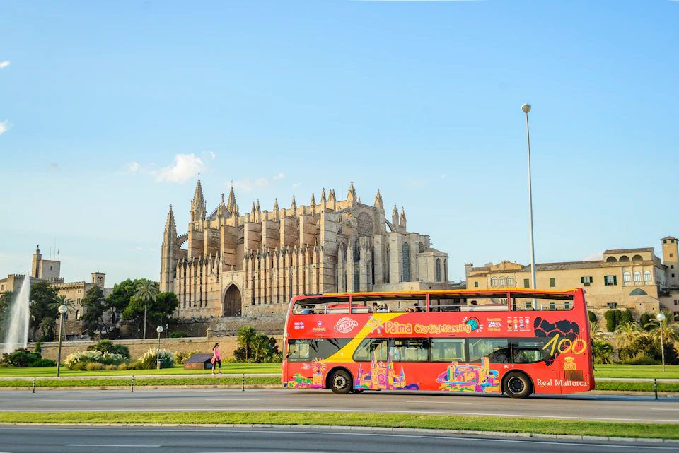 A red double-decker tour bus passes in front of a large Gothic cathedral with multiple spires under a clear blue sky.