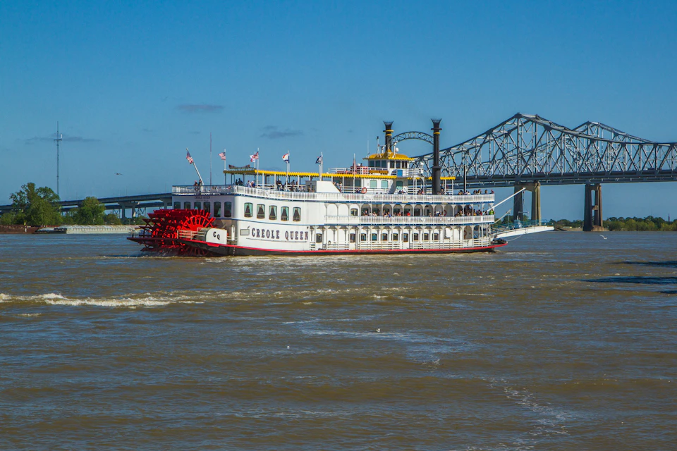 A paddle steamboat named "Creole Queen" on a river with a large bridge in the background under a clear blue sky.