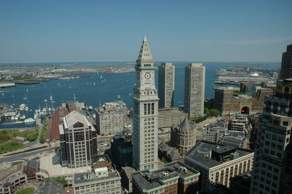 Aerial view of a cityscape featuring a prominent clock tower, high-rise buildings, and a harbor with boats in the background.