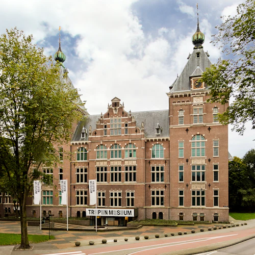 Stor tegelbyggnad märkt "Tropenmuseum" med höga fönster, triangulära gavlar och trädkantad gata i förgrunden.