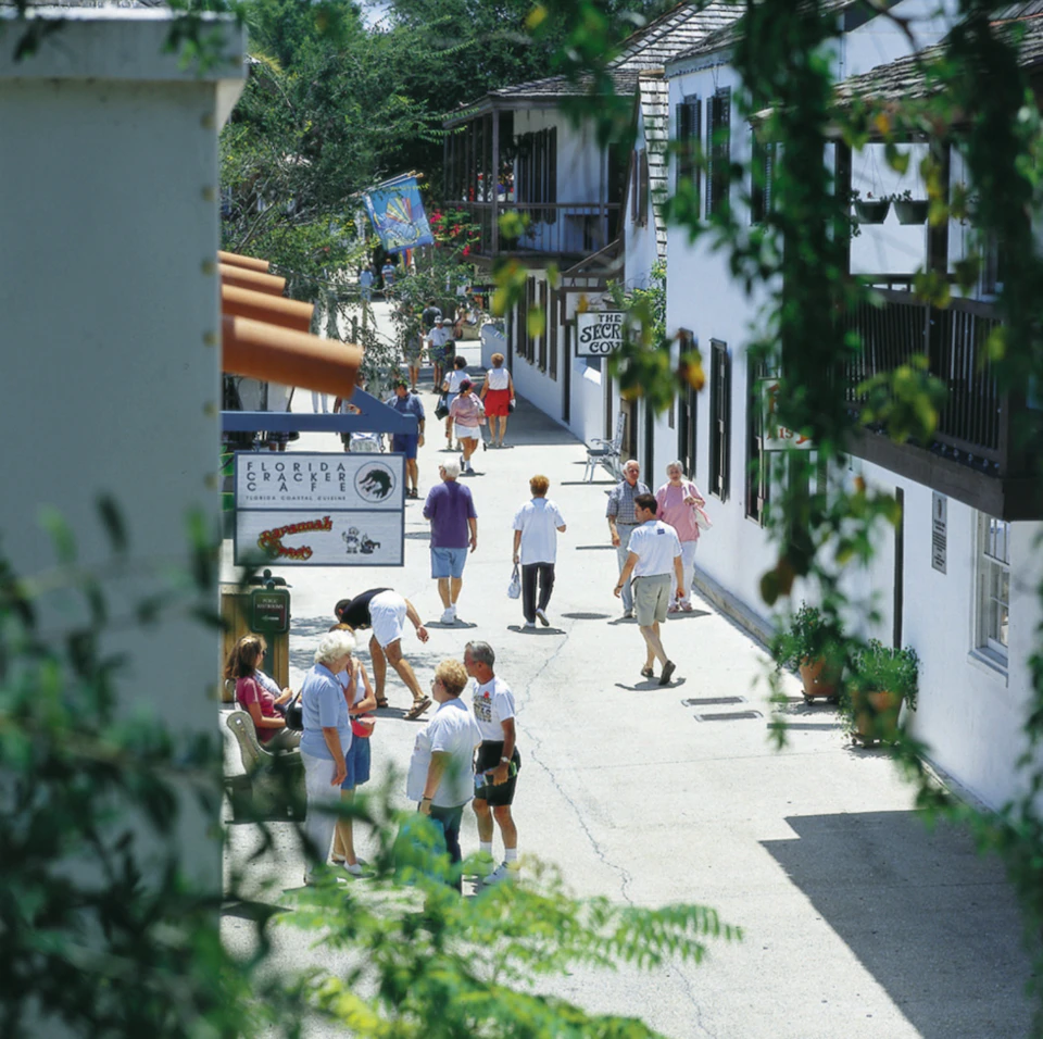People walking and browsing shops on a narrow, sunlit pedestrian street lined with greenery and white buildings.