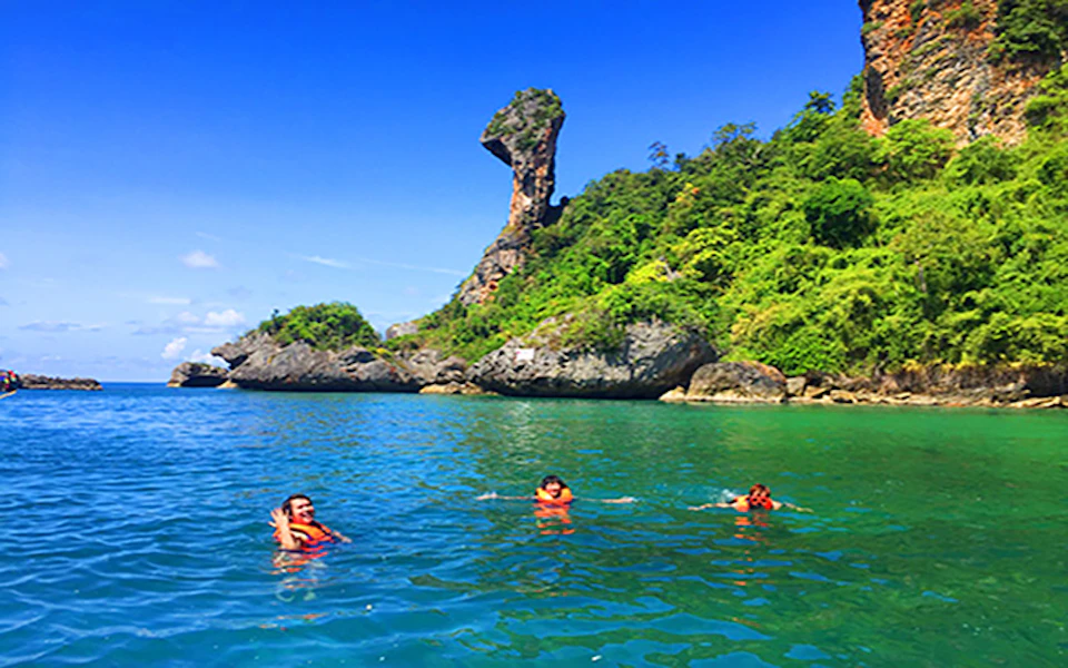 Three people swim in clear blue water near a lush, rocky island with a distinct tall, curved rock formation under a bright blue sky.