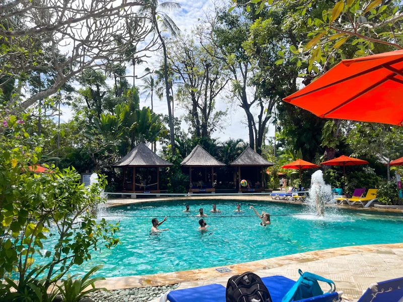 People enjoying a swimming pool surrounded by tropical trees and huts, with red umbrellas and a fountain in the water.