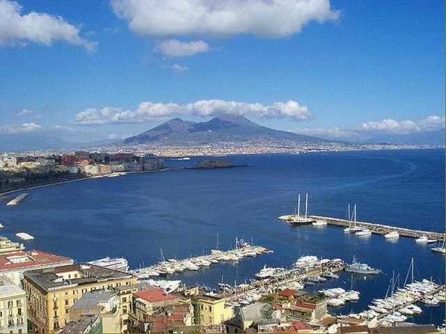 Vue d'une ville côtière avec un port de plaisance, des bateaux amarrés et une grande montagne en arrière-plan, sous un ciel partiellement nuageux.