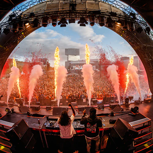 DJs uppträder på scenen med stor publik som jublar, eld- och rökeffekter, under en stor halvcirkelformad struktur.