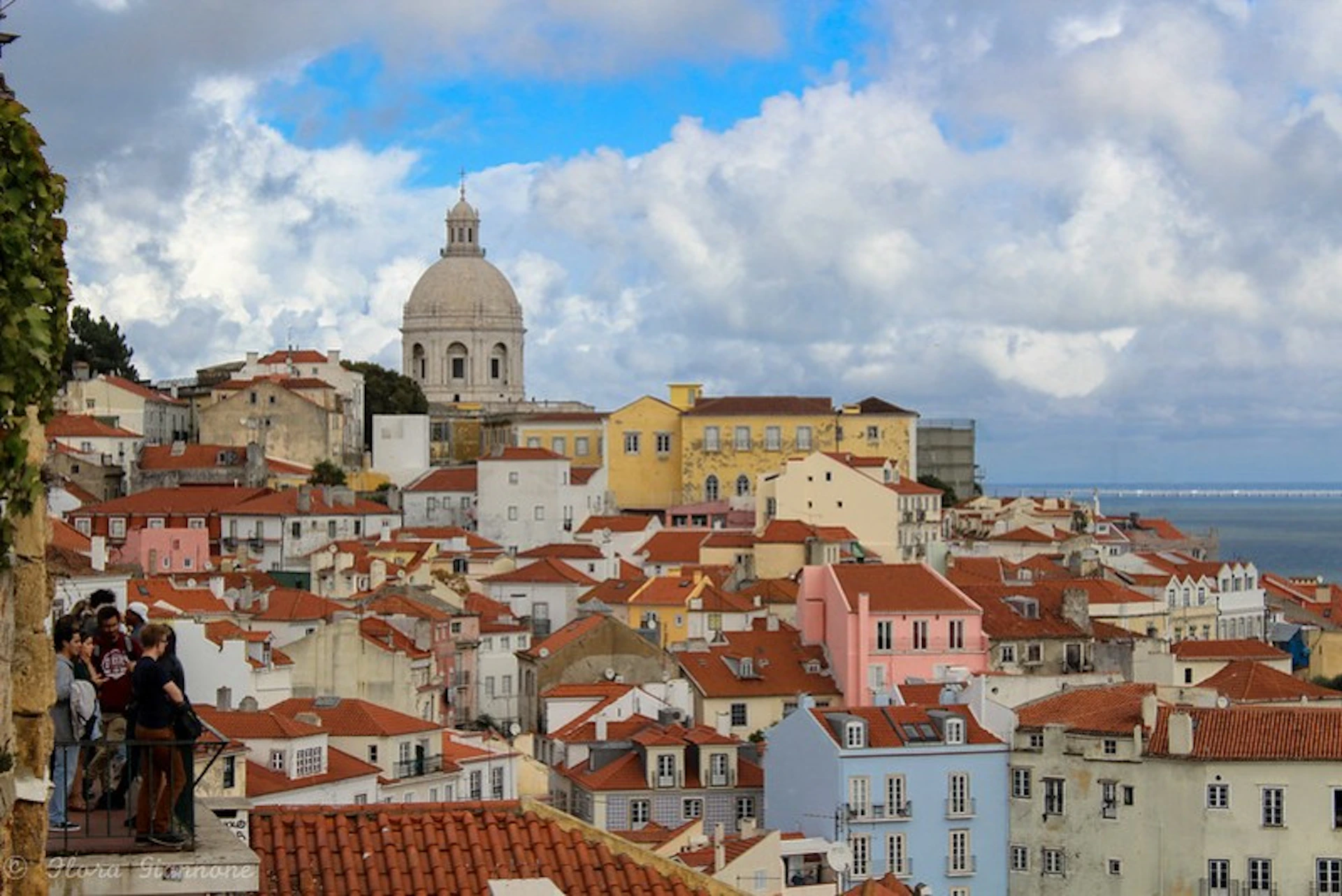 A panoramic view of a town with clustered buildings having red-tiled roofs and a large domed structure in the background under a partly cloudy sky.