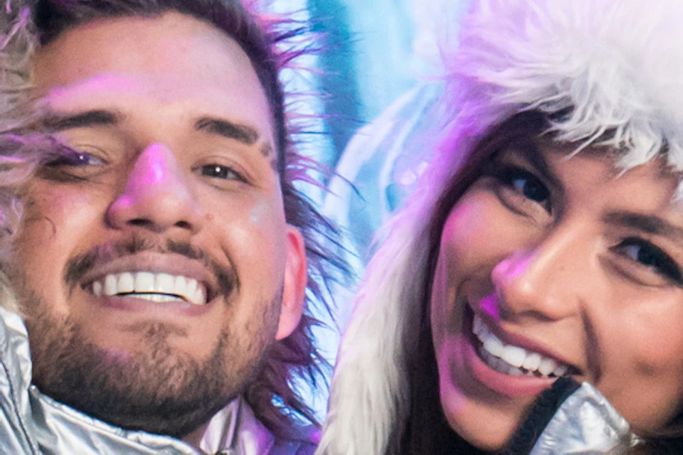 A smiling couple in silver jackets and winter gear pose together in front of ice sculptures with vibrant lighting.
