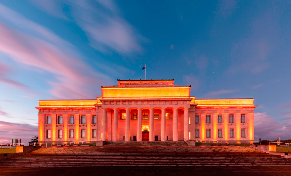 A neoclassical building illuminated in orange light, with columns and steps, under a clear sky with streaks of clouds.