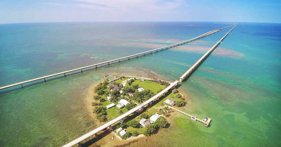 Aerial view of a small island with houses, surrounded by clear blue water and intersected by a long bridge.
