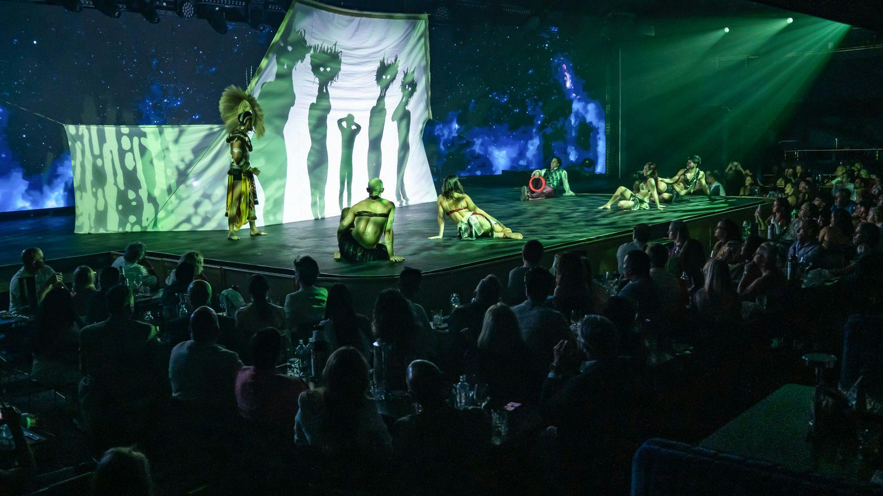 Artistas em trajes tribais num palco com projecções de sombras e um público sentado em primeiro plano.