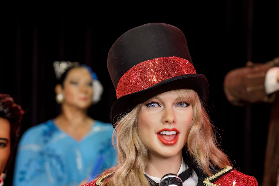Person in a red sequined jacket and top hat holding a microphone, with blurred figures in costume in the background.