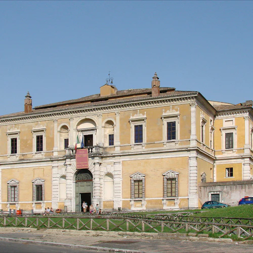Stor gul historisk byggnad med välvd ingång, stendetaljer och spärrade fönster. Få människor och parkerade bilar utanför. Klar himmel.