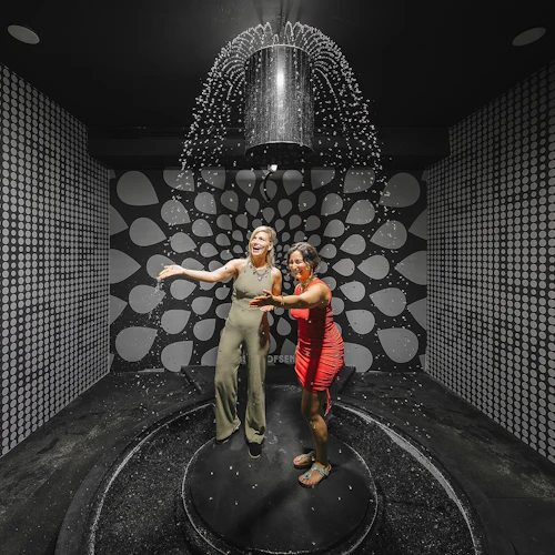 Två kvinnor står på en plattform under en vattenfunktion, omgiven av en mönstrad svartvit vägg, leende och interagerande.