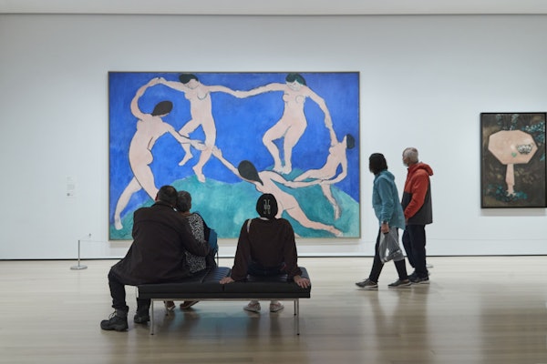 Fem personer ser et stort maleri af dansende figurer i et moderne kunstgalleri. To sidder på en bænk, mens tre står.