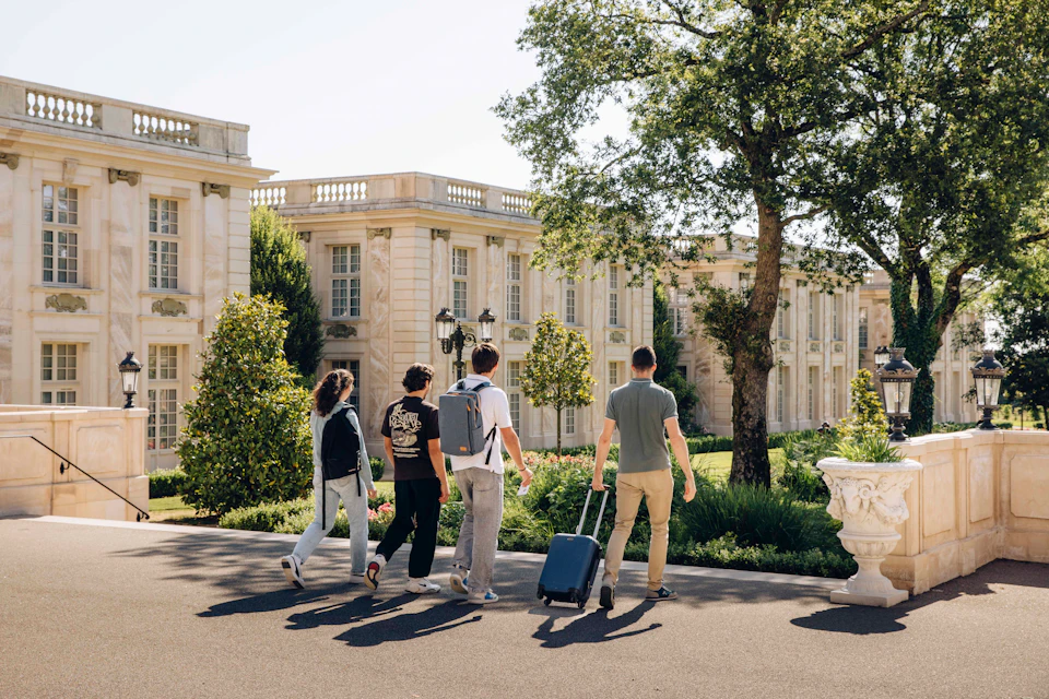 Four people walking towards a classical-style building with luggage, surrounded by trees and greenery on a sunny day.