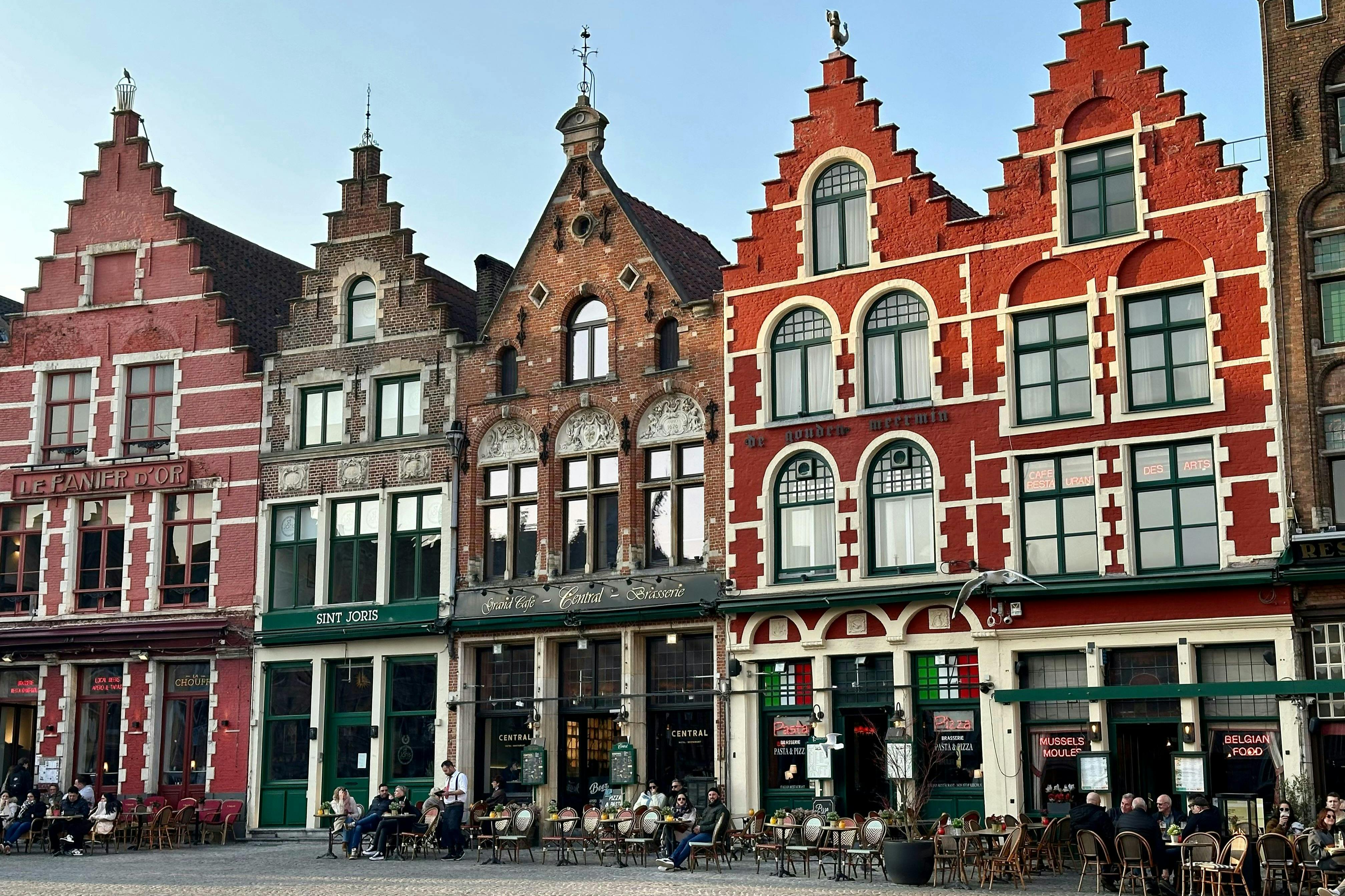 People sit at outdoor café tables in front of three historic, gabled buildings with ornate facades and large windows.