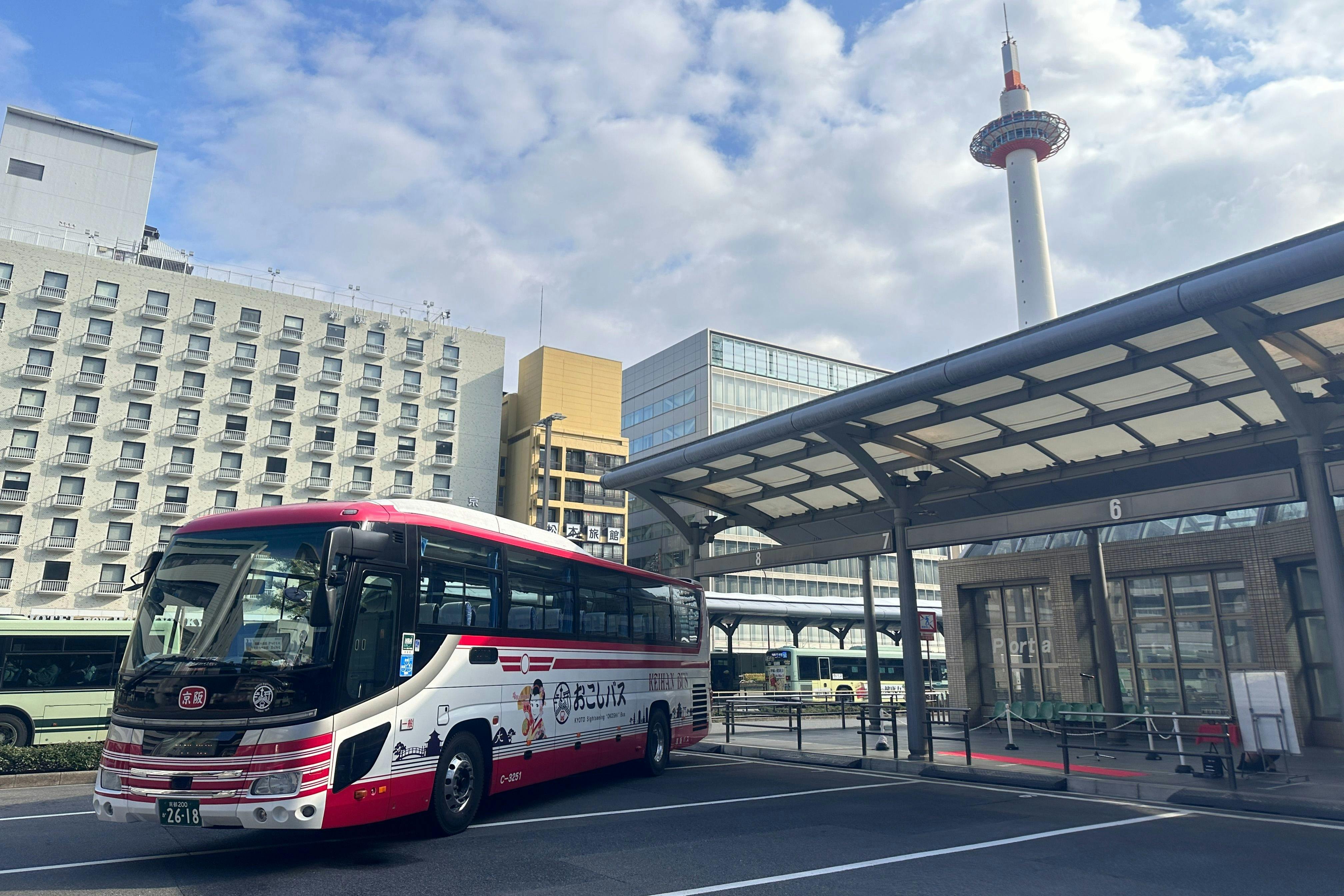 Ένα κόκκινο-άσπρο λεωφορείο σταθμευμένο σε ένα σταθμό λεωφορείων με σύγχρονα κτίρια και τον Πύργο του Κιότο στο παρασκήνιο κάτω από έναν εν μέρει συννεφιασμένο ουρανό.