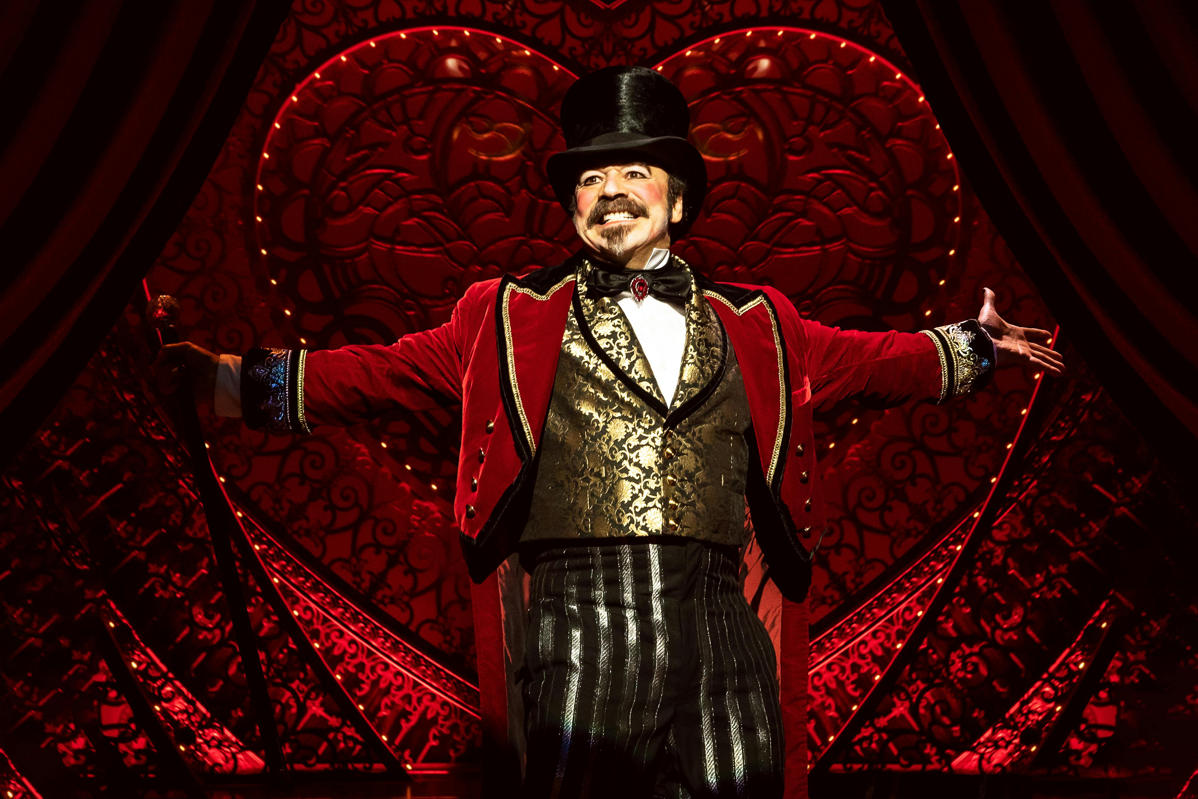 Man in a red and gold ornate costume with a top hat stands on stage with arms outstretched, a red heart-shaped backdrop behind him.