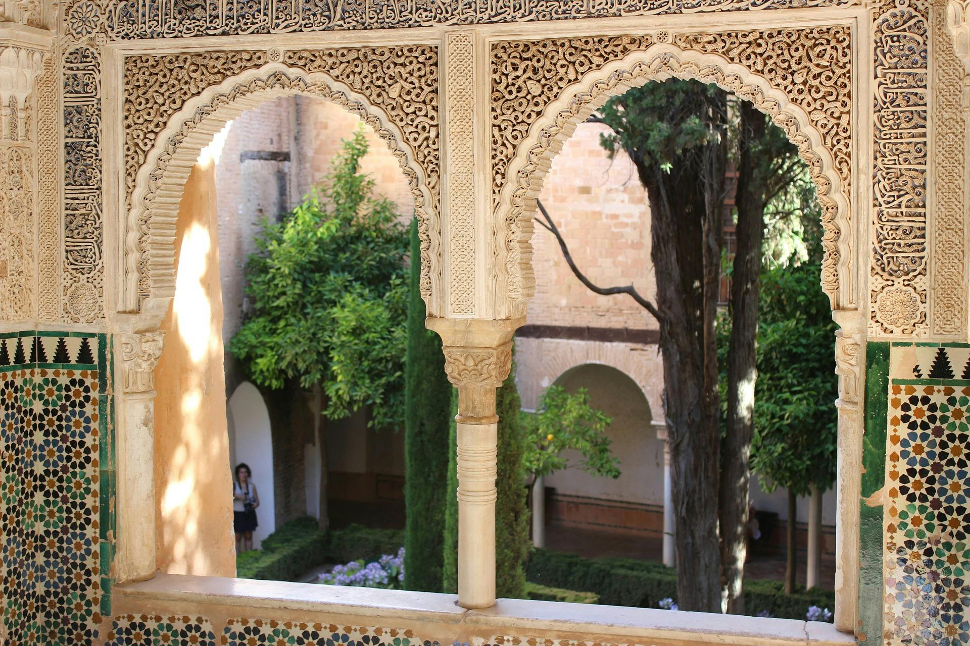 Ornate stone arches frame a courtyard garden with trees and shrubs. A person stands near the wall in the background.