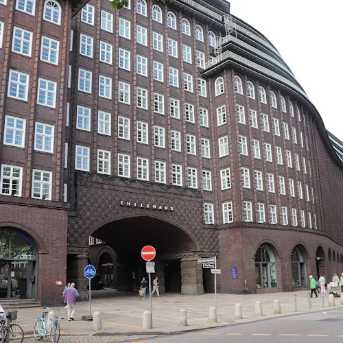Tegelbyggnad med många fönster, välvd entré märkt "Chilehaus", cyklar, fotgängare och gatuskyltar i förgrunden.