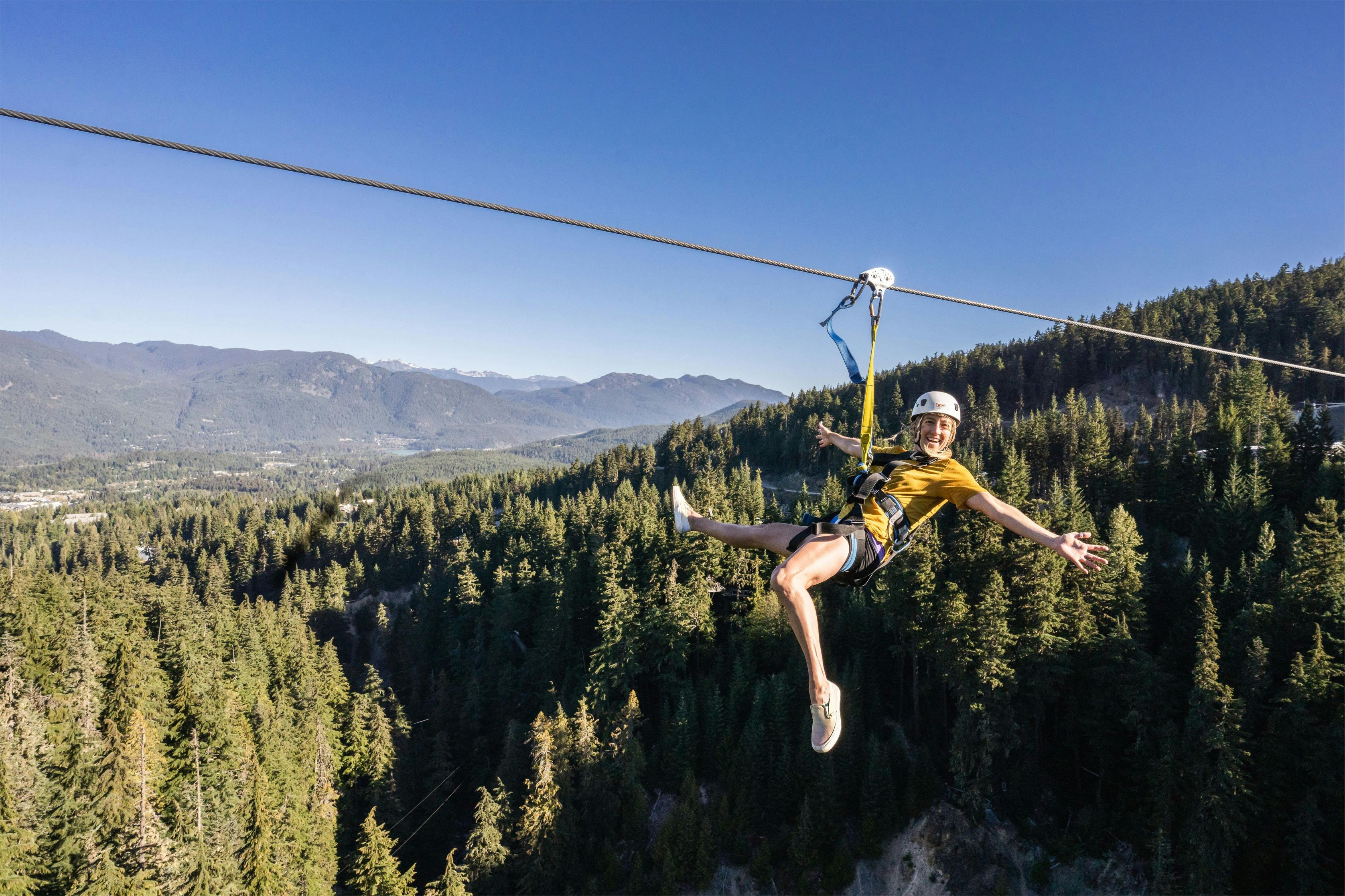 노란색 셔츠와 헬멧을 쓴 사람이 산을 배경으로 빽빽한 숲 위에 지퍼 라이닝을 즐기고 있습니다.