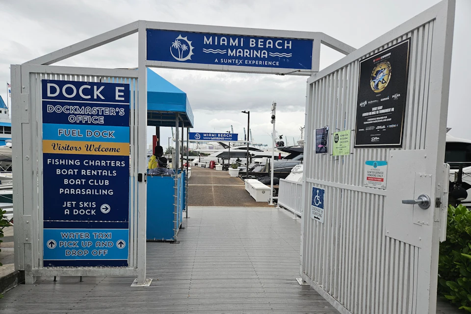 Entrance gate to Miami Beach Marina with signs for various services, boats docked in the background, and a cloudy sky overhead.