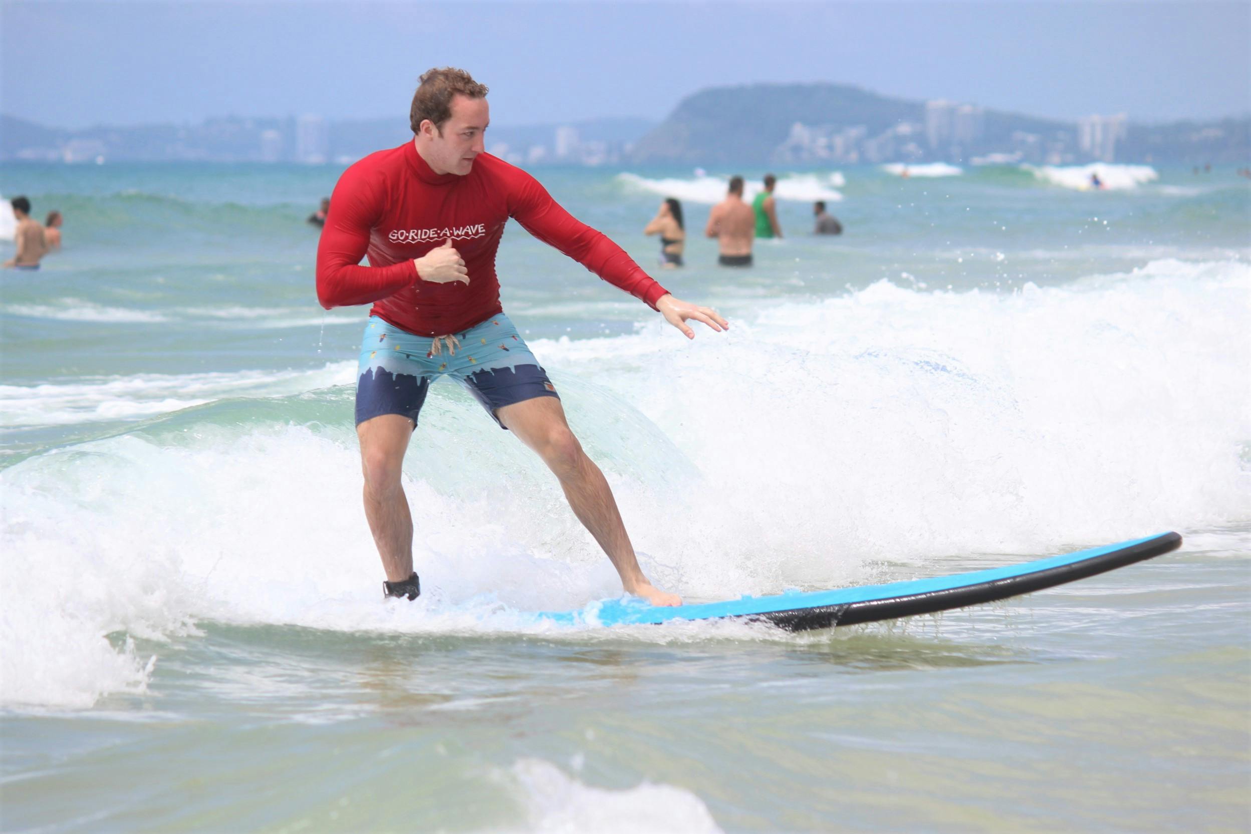 一名男子在海滩上冲浪，身穿红色衬衫和蓝色短裤，其他人则在后台游泳。