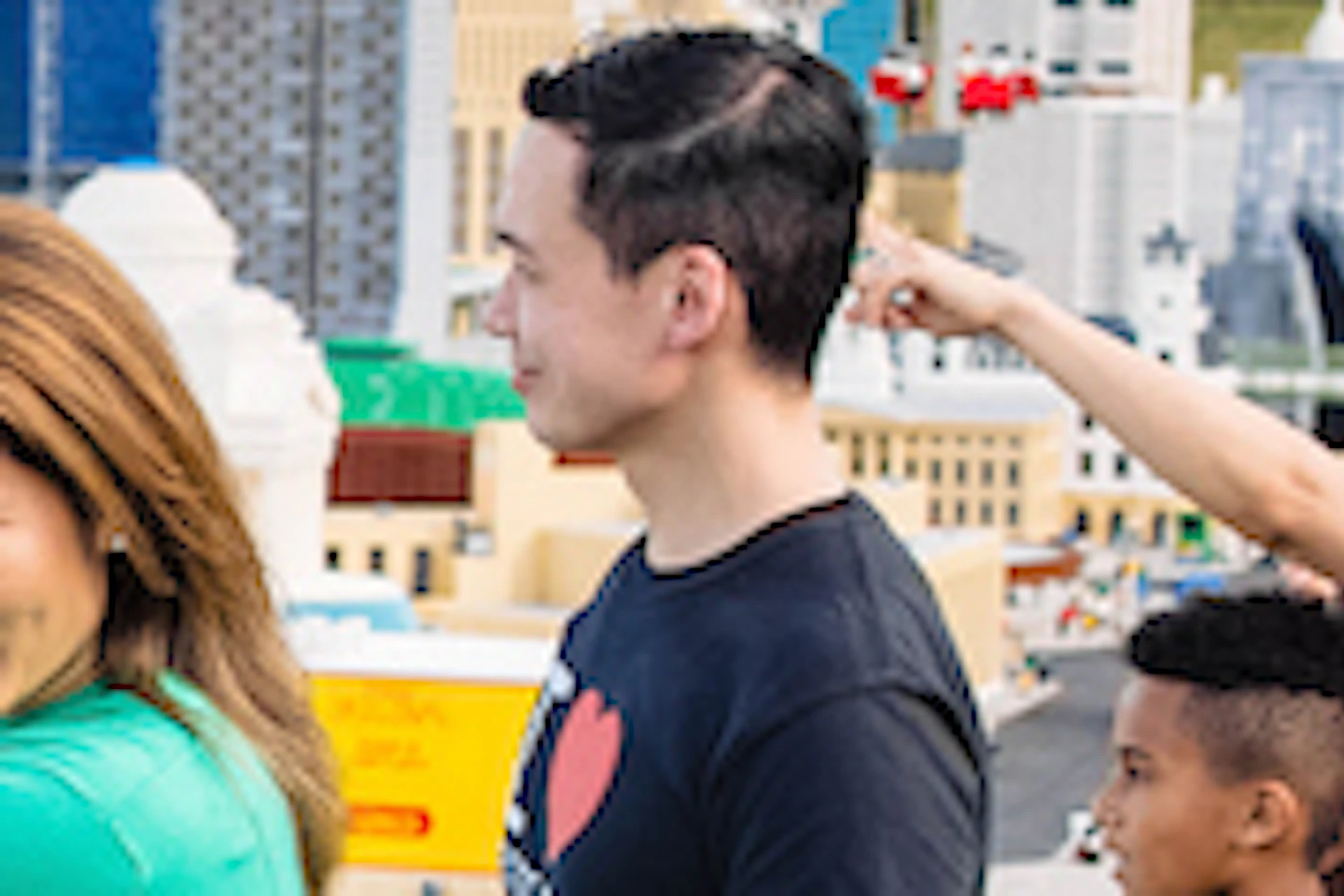A family poses near LEGO character sculptures and the entrance sign to LEGOLAND New York on a sunny day.