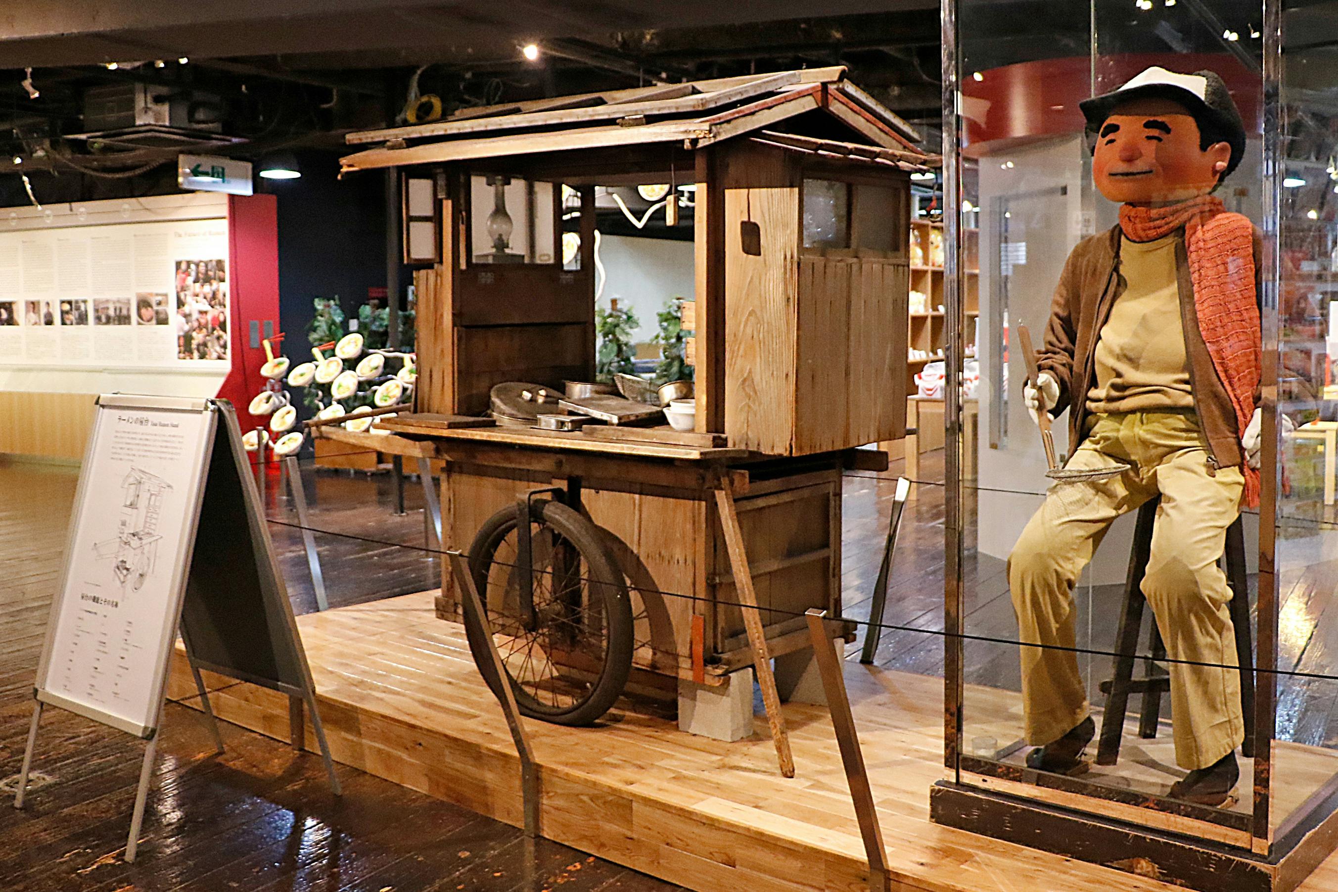 A museum display featuring a wooden street cart and a mannequin seated in a glass case, dressed in casual attire.