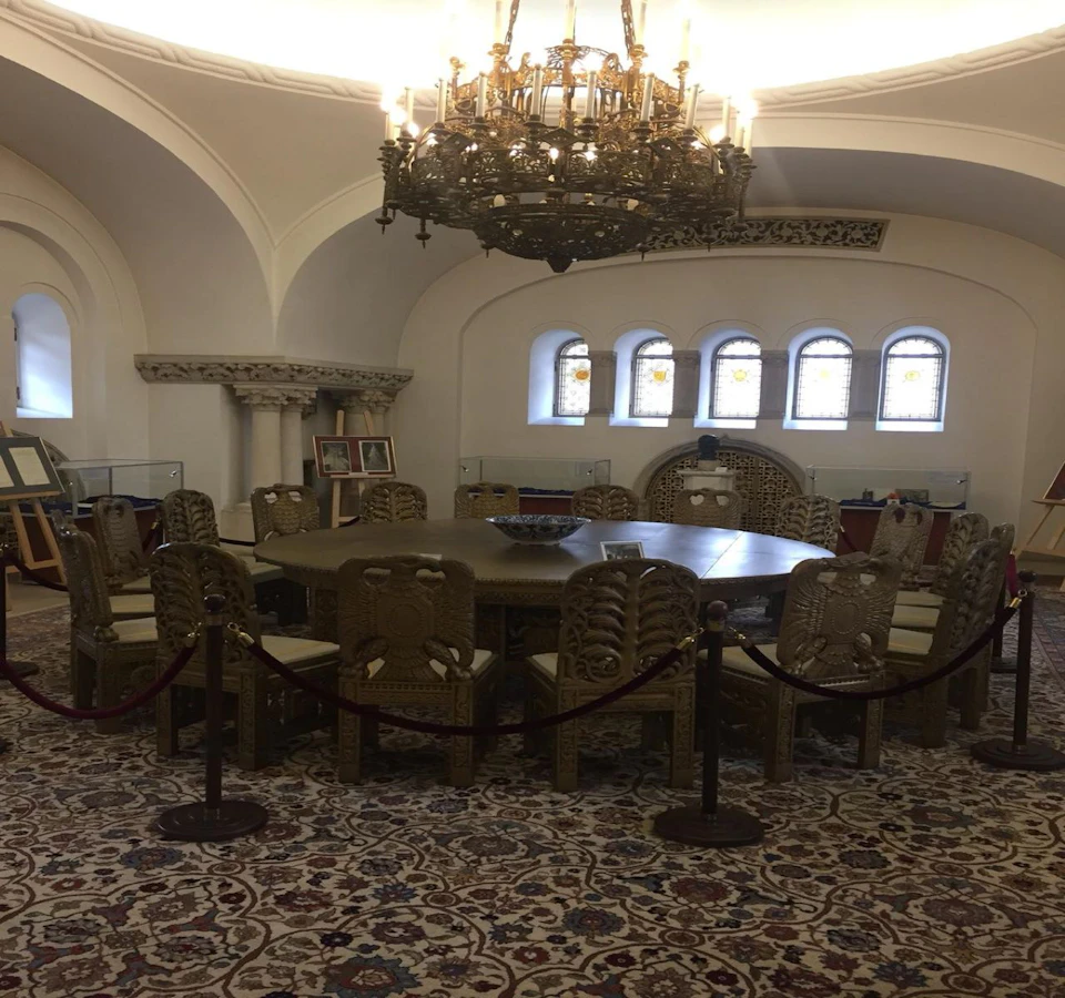 Ornate meeting room with a round table and carved wooden chairs, surrounded by stained glass windows and a large chandelier above.
